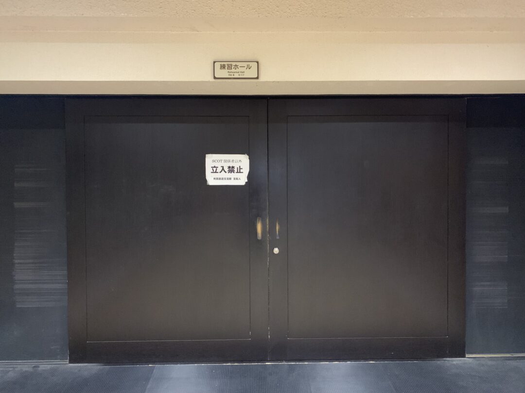 una porta a due battenti nera chiusa con un cartello che in giapponese indica la sala come proprietà della SCOT e un altro cartello sopra il muro che dichiara in inglese e giapponese "sala prove"