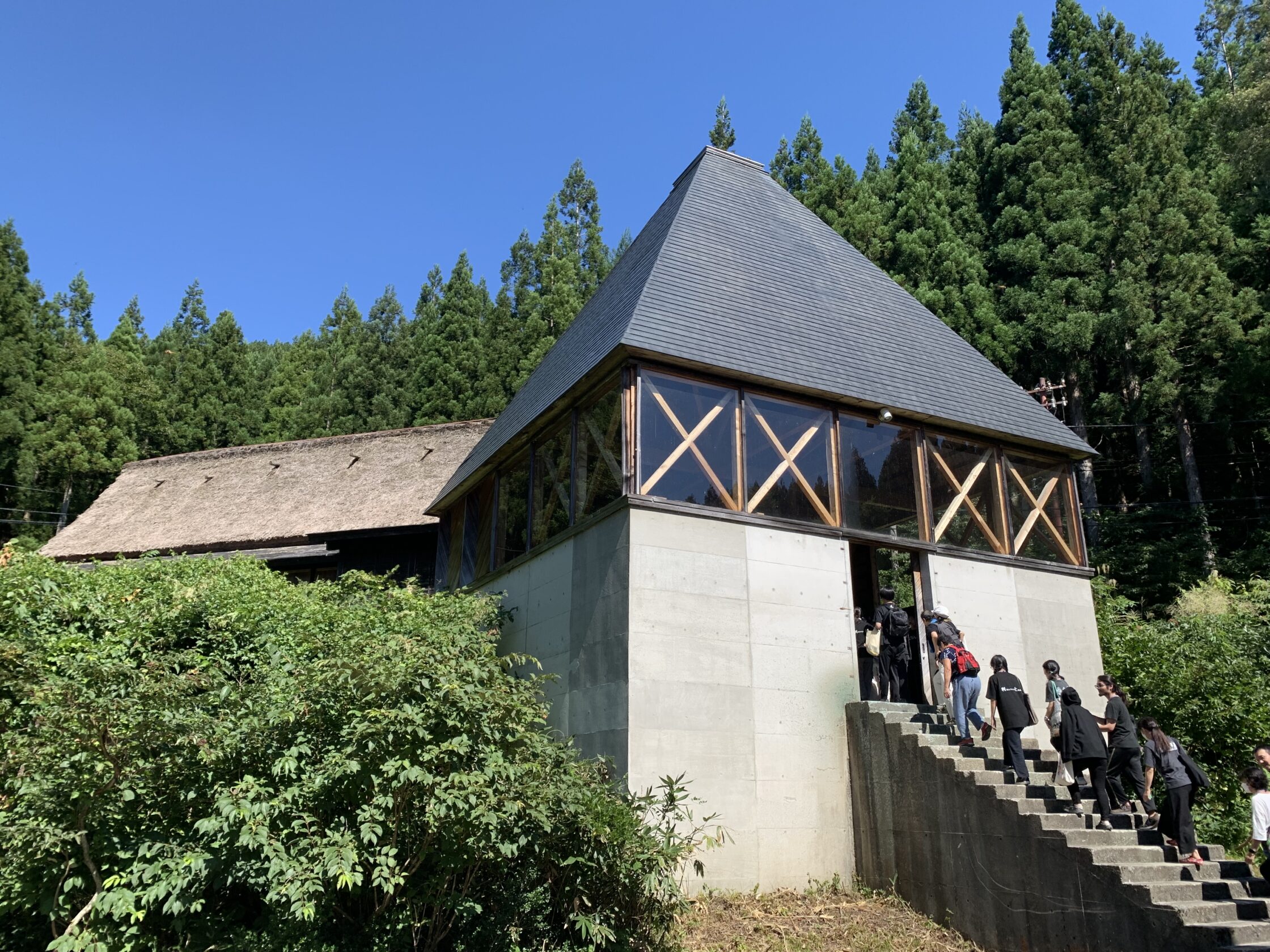 un edificio a forma di parallelepipedo in cemento, sormontato da un tetto piramidale che insiste su una cornice di finestre. sullo sfondo il tetto di una fattoria di montagna tradizionale giapponese